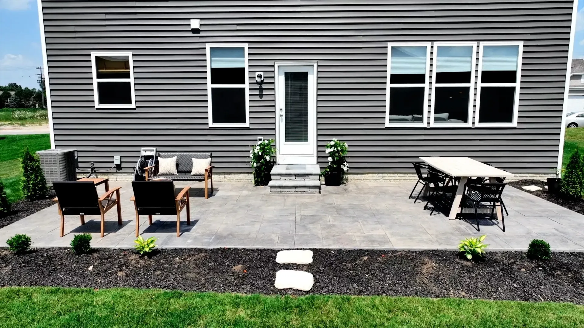 Backyard patio with seating area, dining table, and dark gray siding. Black chairs, a bench, and potted plants are visible.