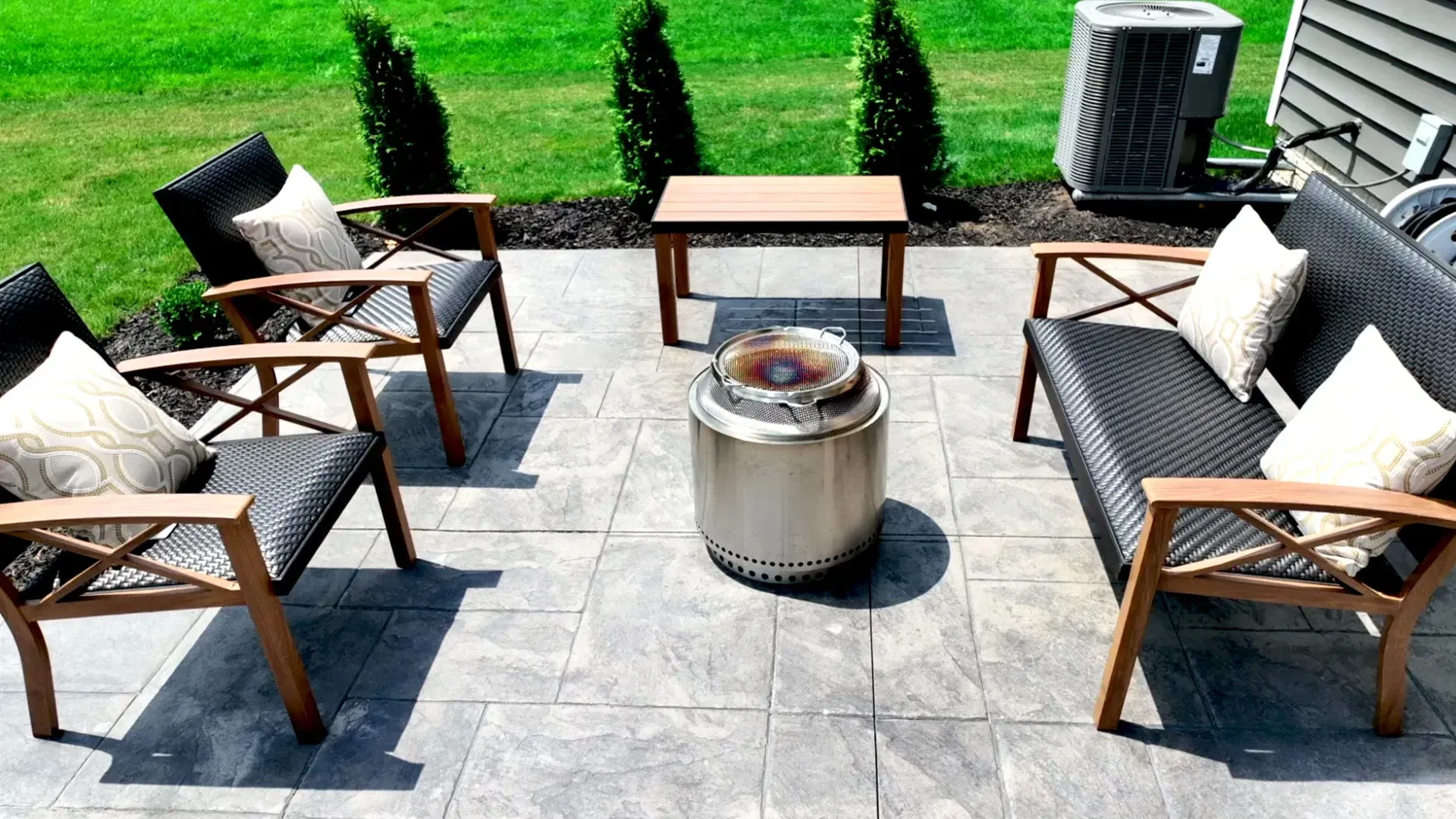 Patio with seating arranged around a stainless steel fire pit. Includes four chairs, a sofa, a small table, and greenery.