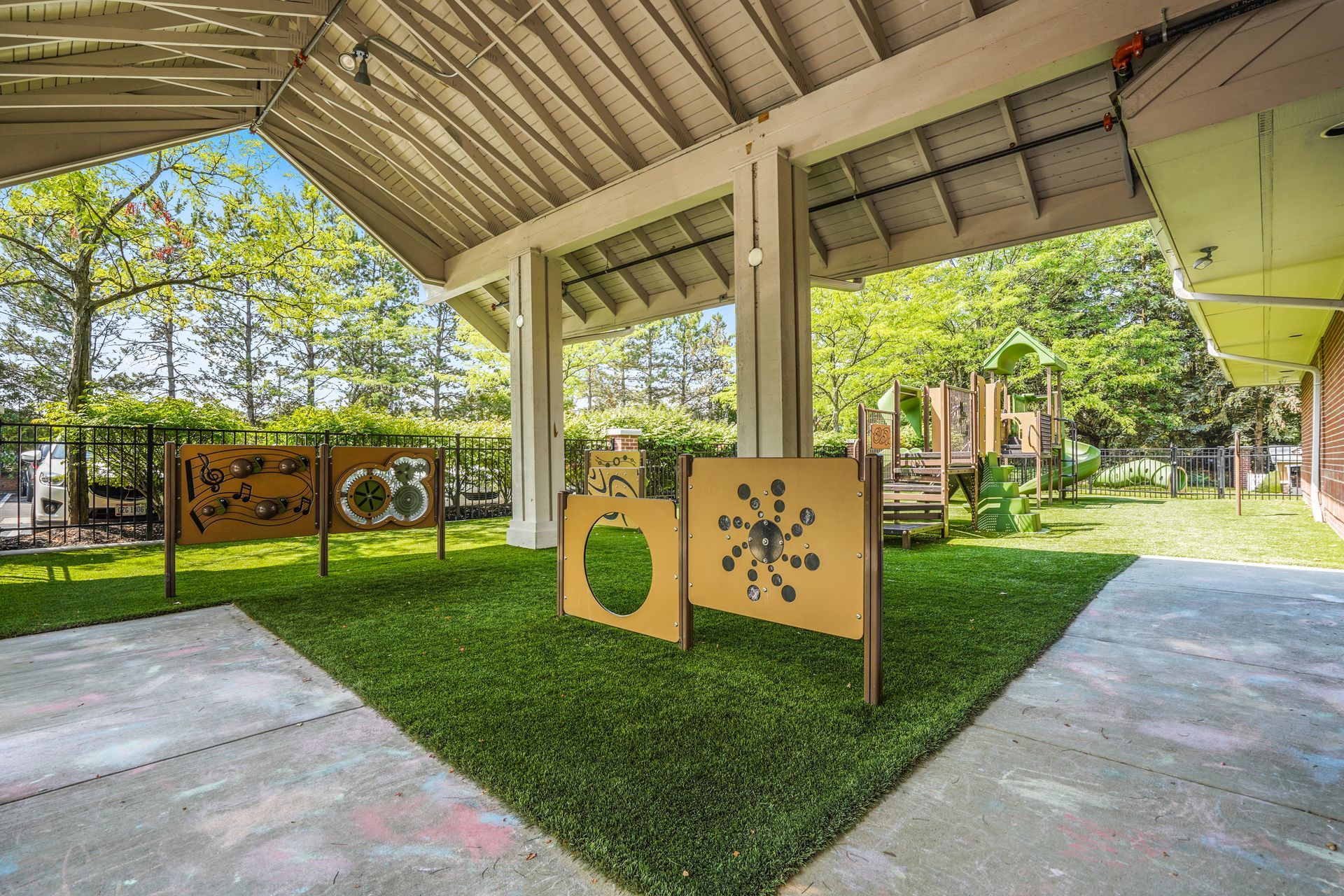 A partially covered outdoor play area with artificial turf and educational play panels. Trees and a wooden playset are in the background.