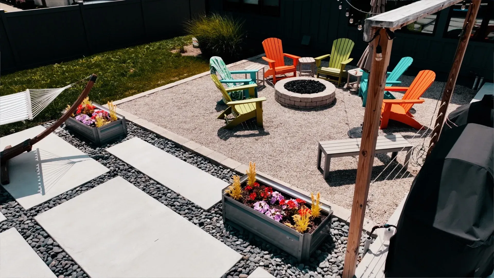 Backyard patio with colorful Adirondack chairs around a fire pit. Concrete pavers and a small garden with flower boxes.