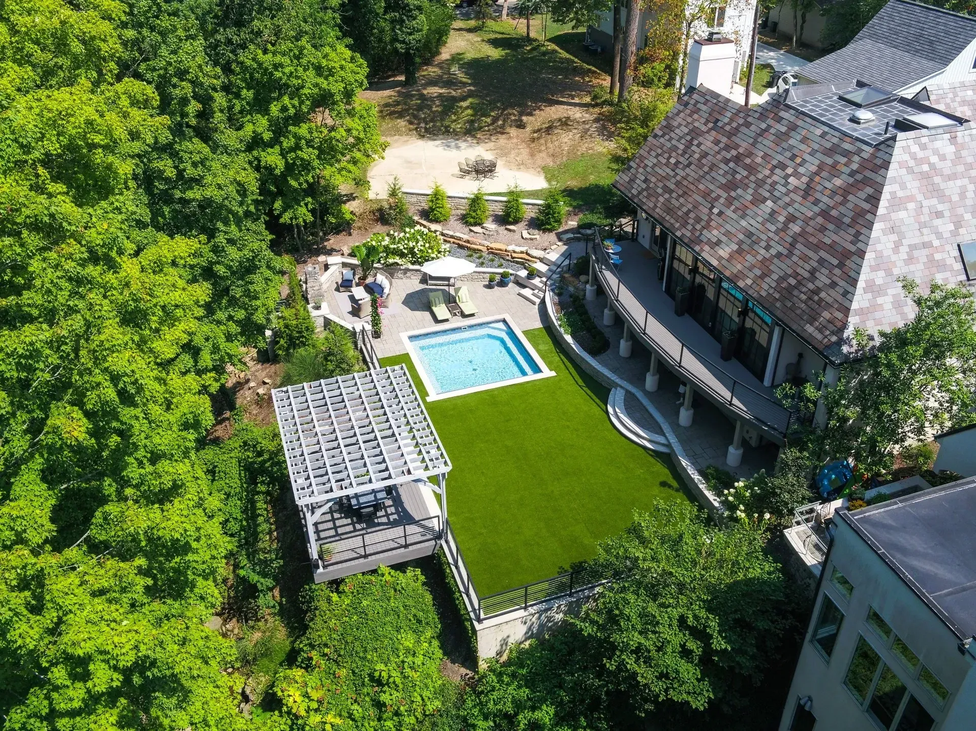 Aerial view of a modern house with a pool, patio, and pergola surrounded by green lawn and trees.