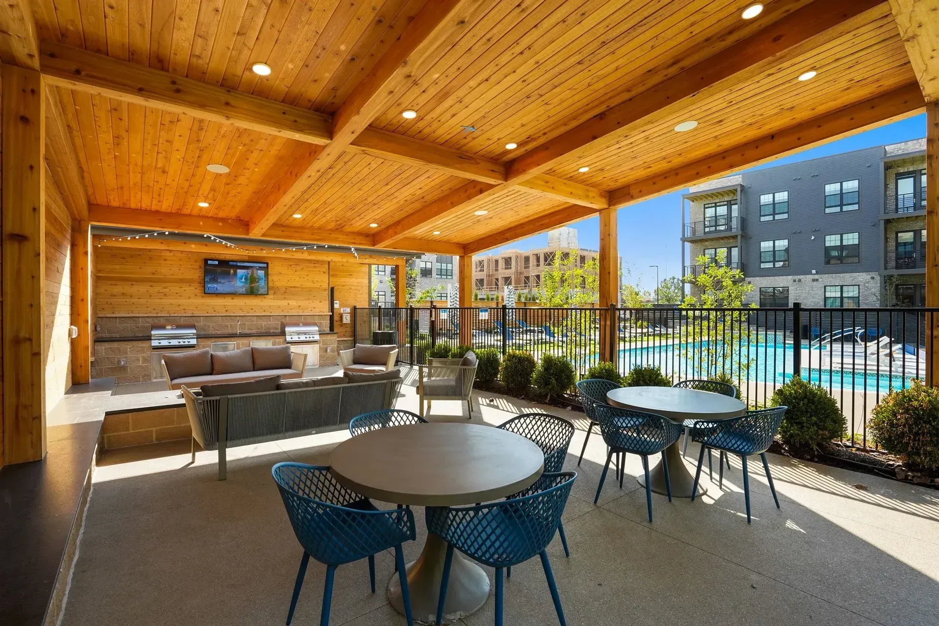 Outdoor patio with seating, a grill, and a TV. The area is next to a pool and apartment buildings on a sunny day.