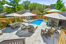Backyard patio with pool, lounge chairs, picnic table, and umbrellas under a sunny sky. Green trees and homes in the background.