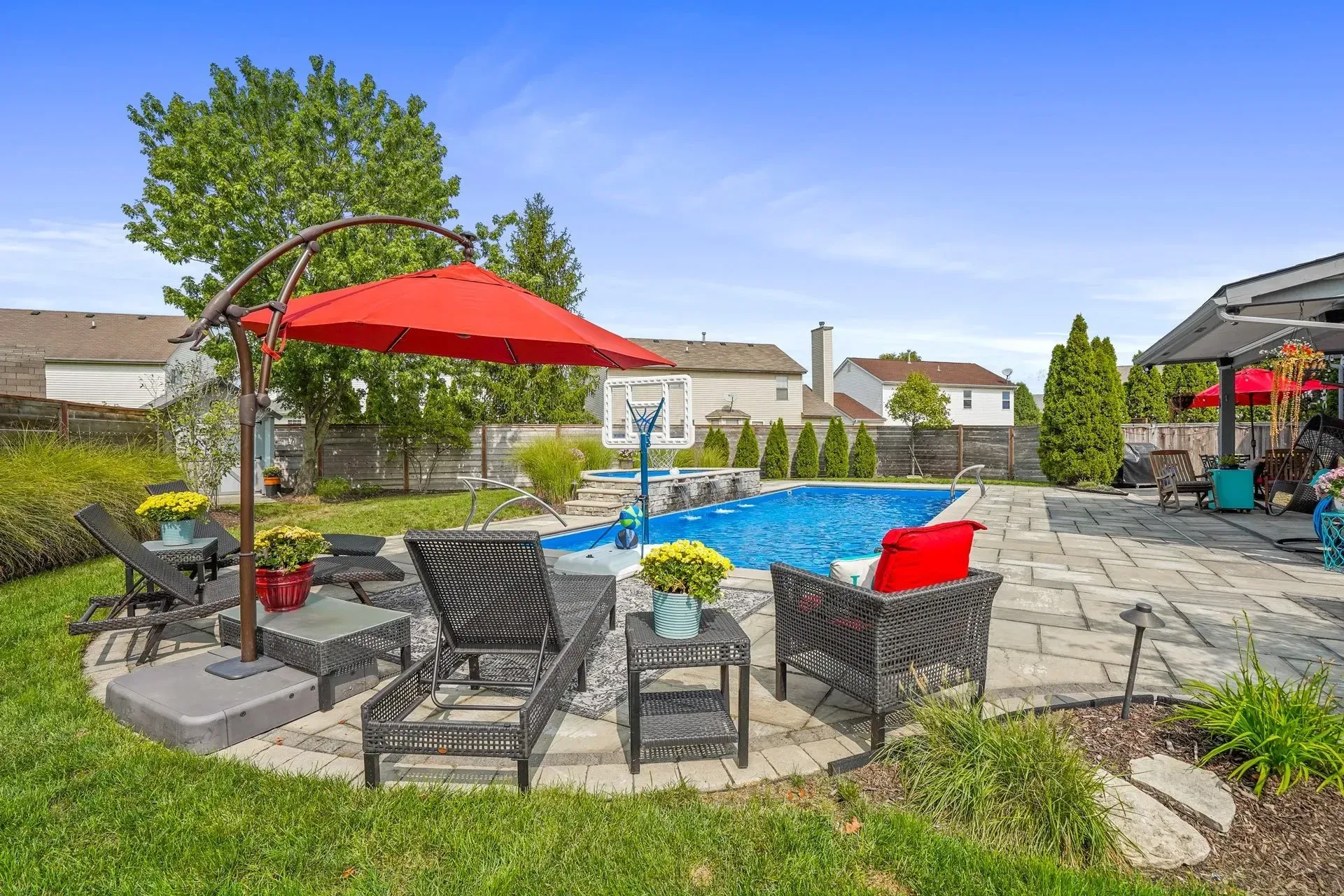Backyard with a pool, lounge chairs, red umbrella, and brick patio. Houses and trees are in the background, under a blue sky.