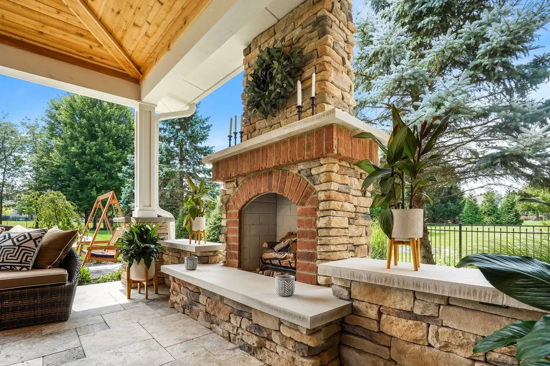 Outdoor patio with stone fireplace and wood-paneled ceiling.  Green foliage surrounds the space with a view of trees and a fence.