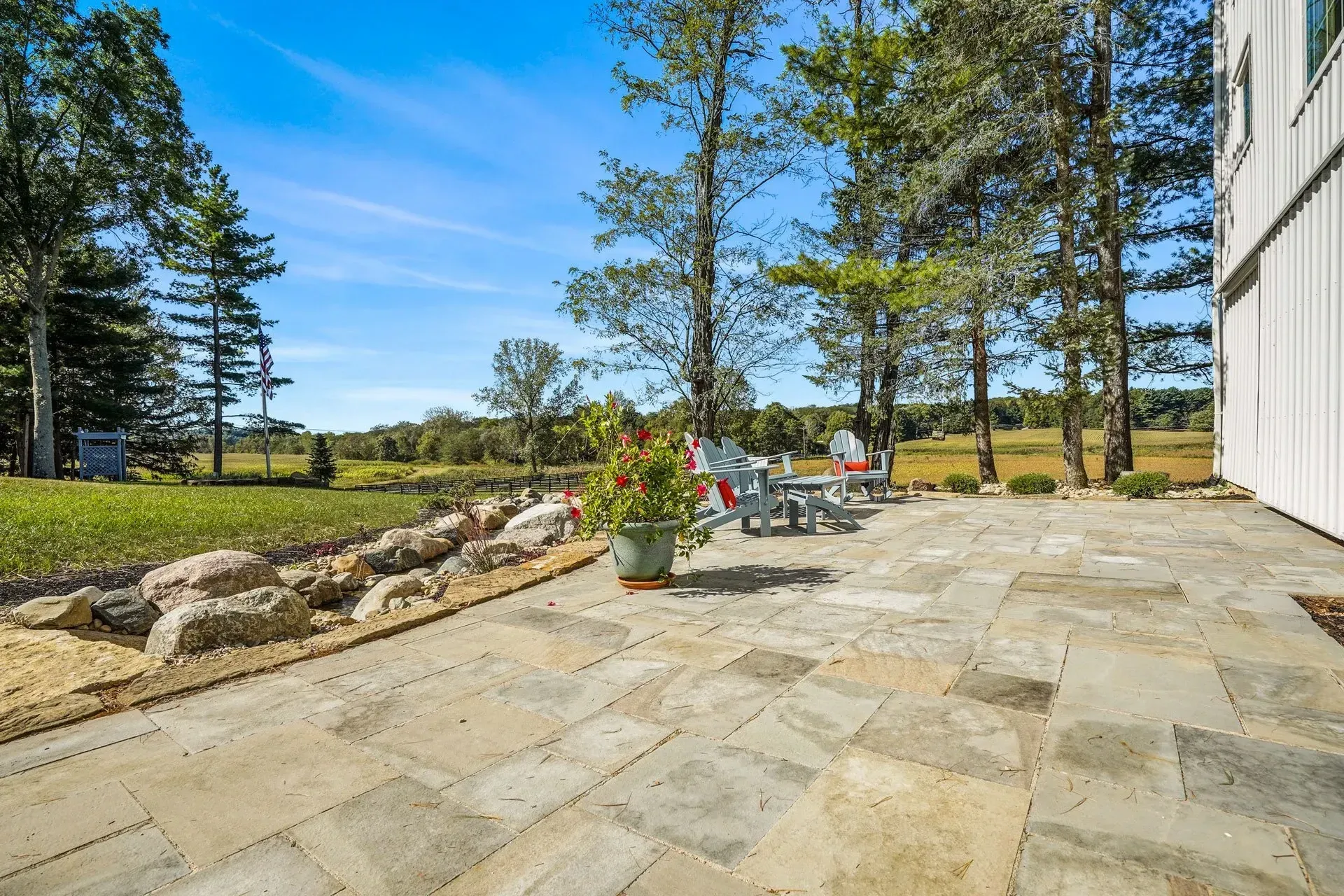 Stone patio with outdoor furniture, a potted plant, and a rock garden next to a white building, with a grassy field and trees in the background.