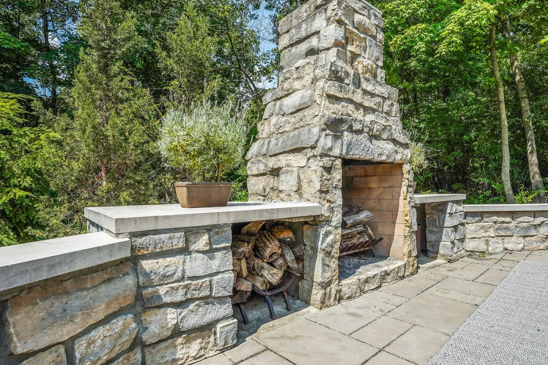 Stone outdoor fireplace with wood storage, on a stone patio, backed by lush greenery. A potted plant rests on the nearby ledge.