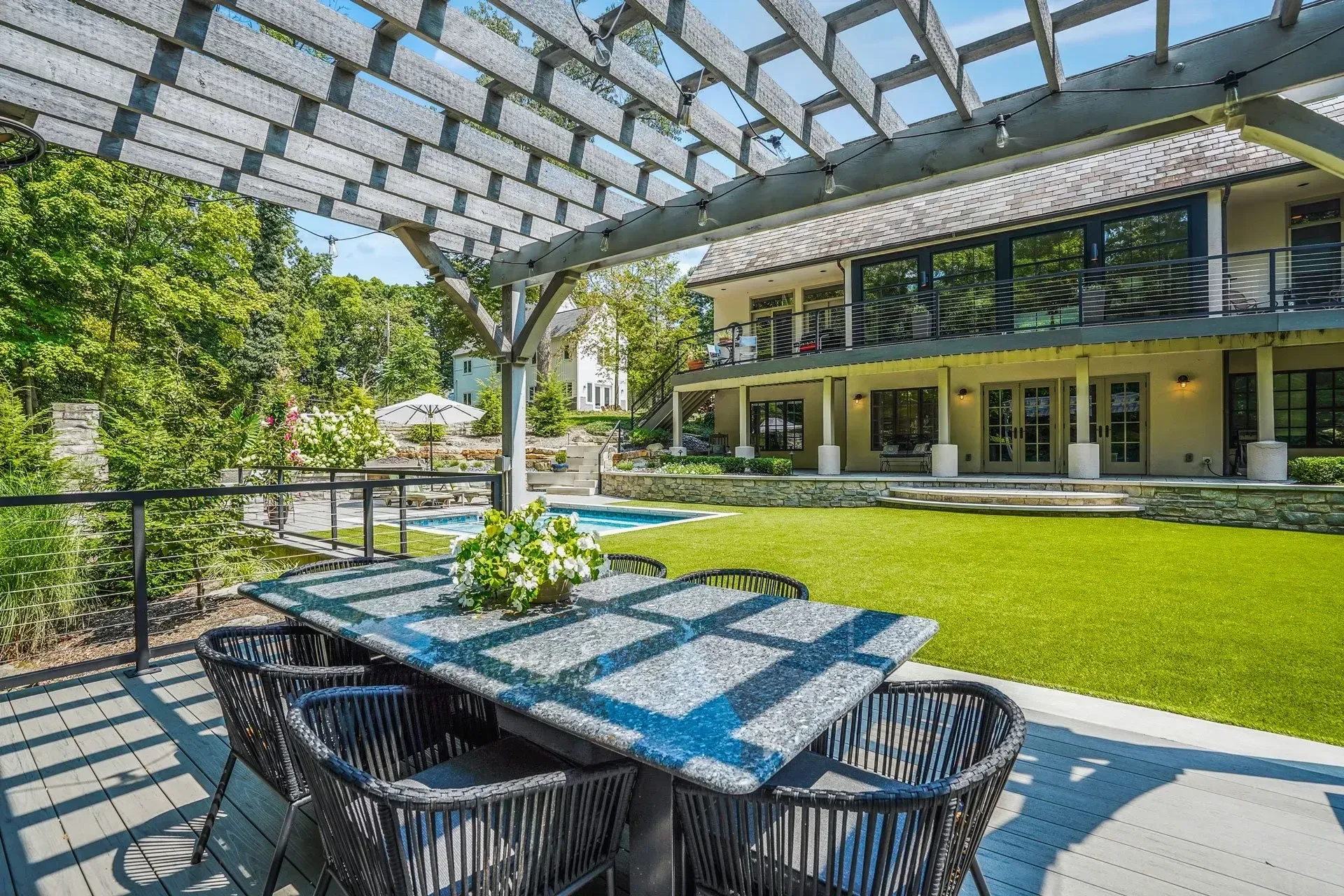 Patio with table and chairs under a wooden pergola, overlooking a backyard with a pool and a two-story house.