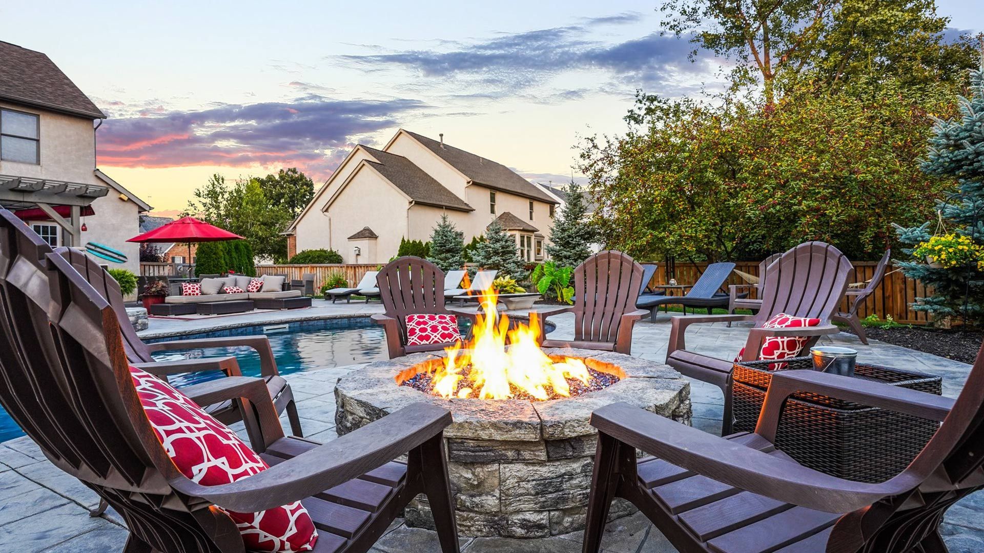 Fire pit surrounded by Adirondack chairs on a patio near a pool, with houses and trees in the background at sunset.