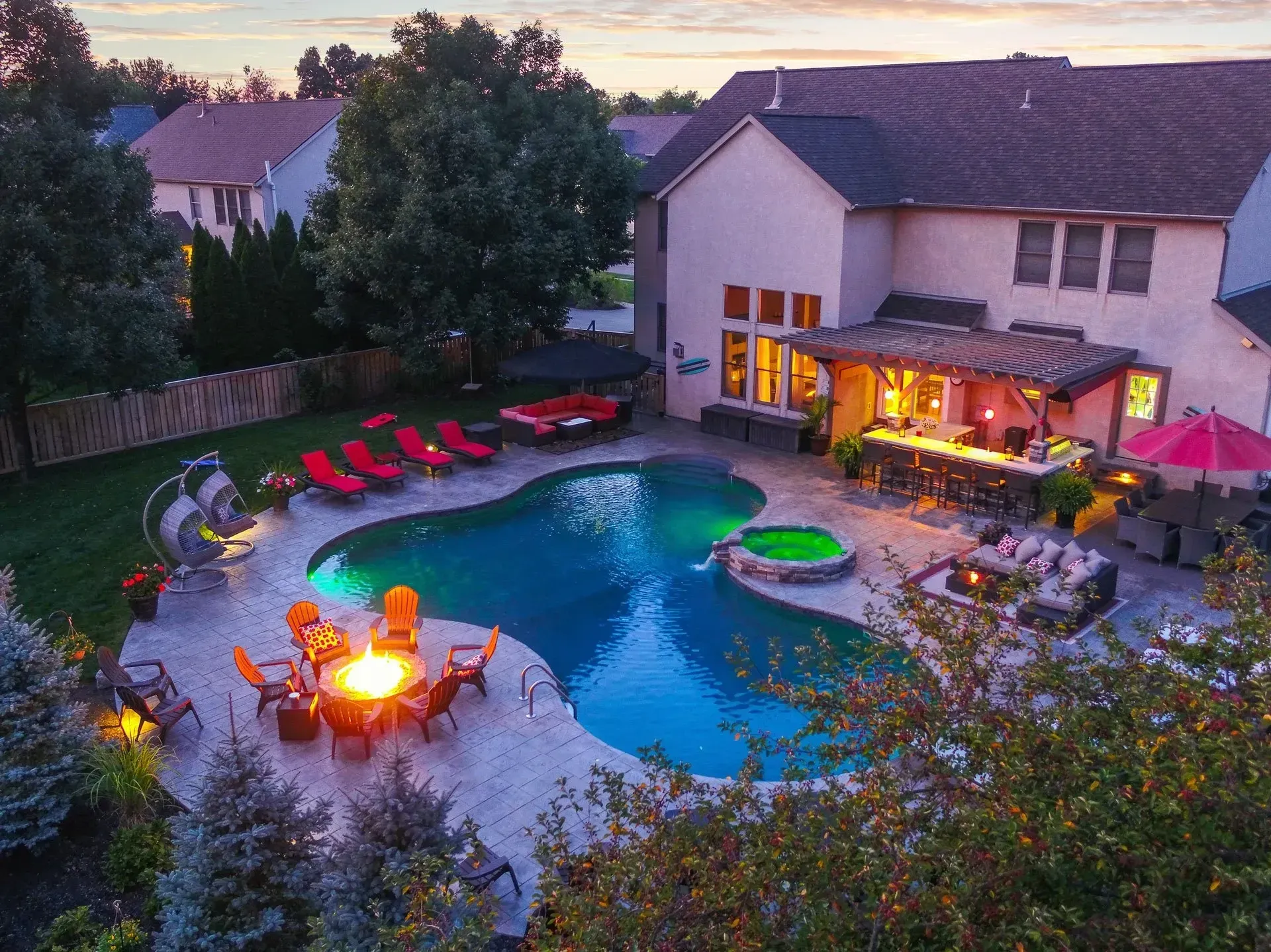 Backyard with pool, fire pit, and bar area illuminated at dusk. Houses in the background.