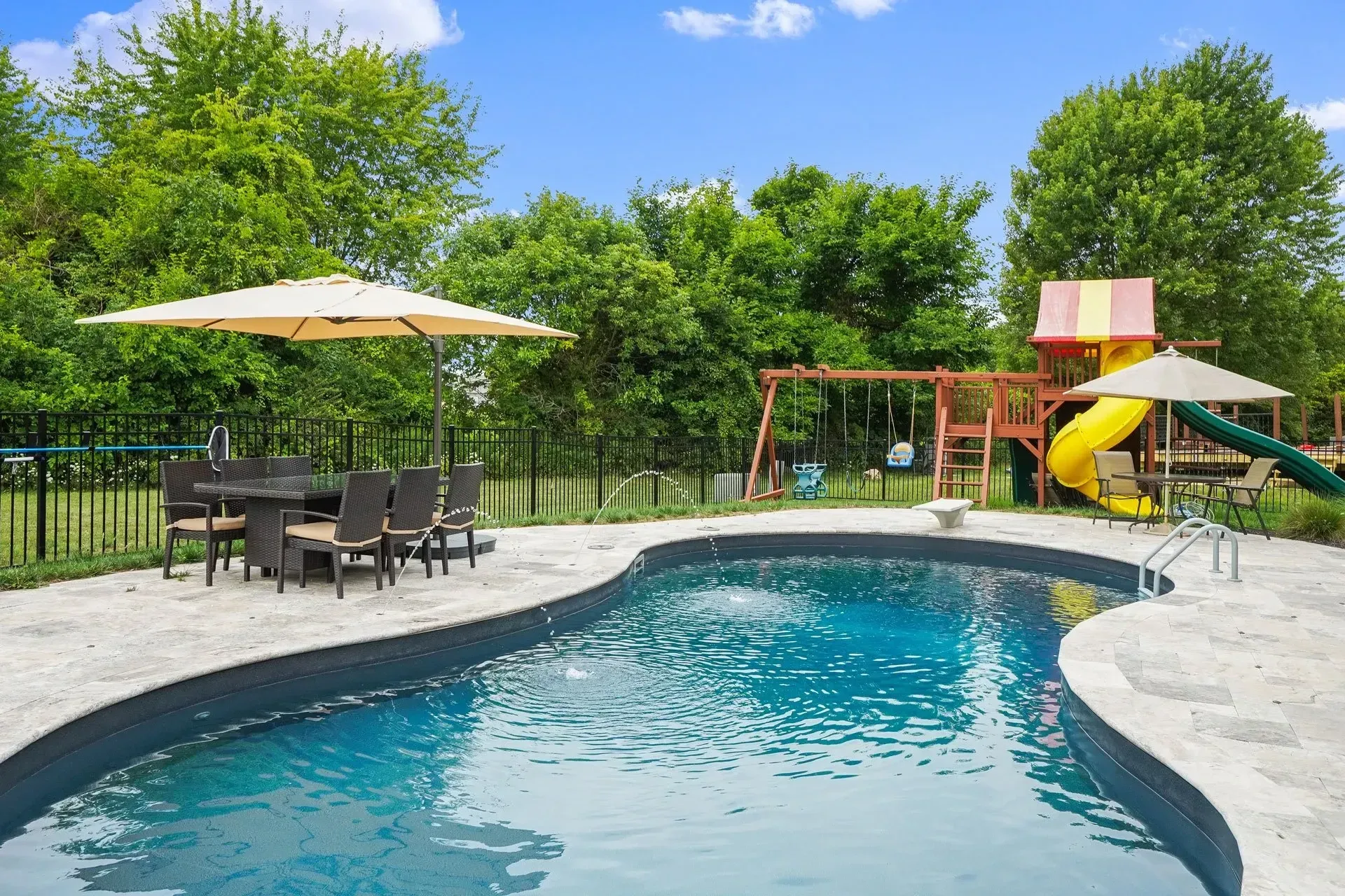 A backyard with a swimming pool, patio furniture, and a playground under a blue sky. Lush green trees surround the area.