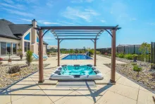 A hot tub under a pergola in a backyard with a pool, blue sky, and a large house.