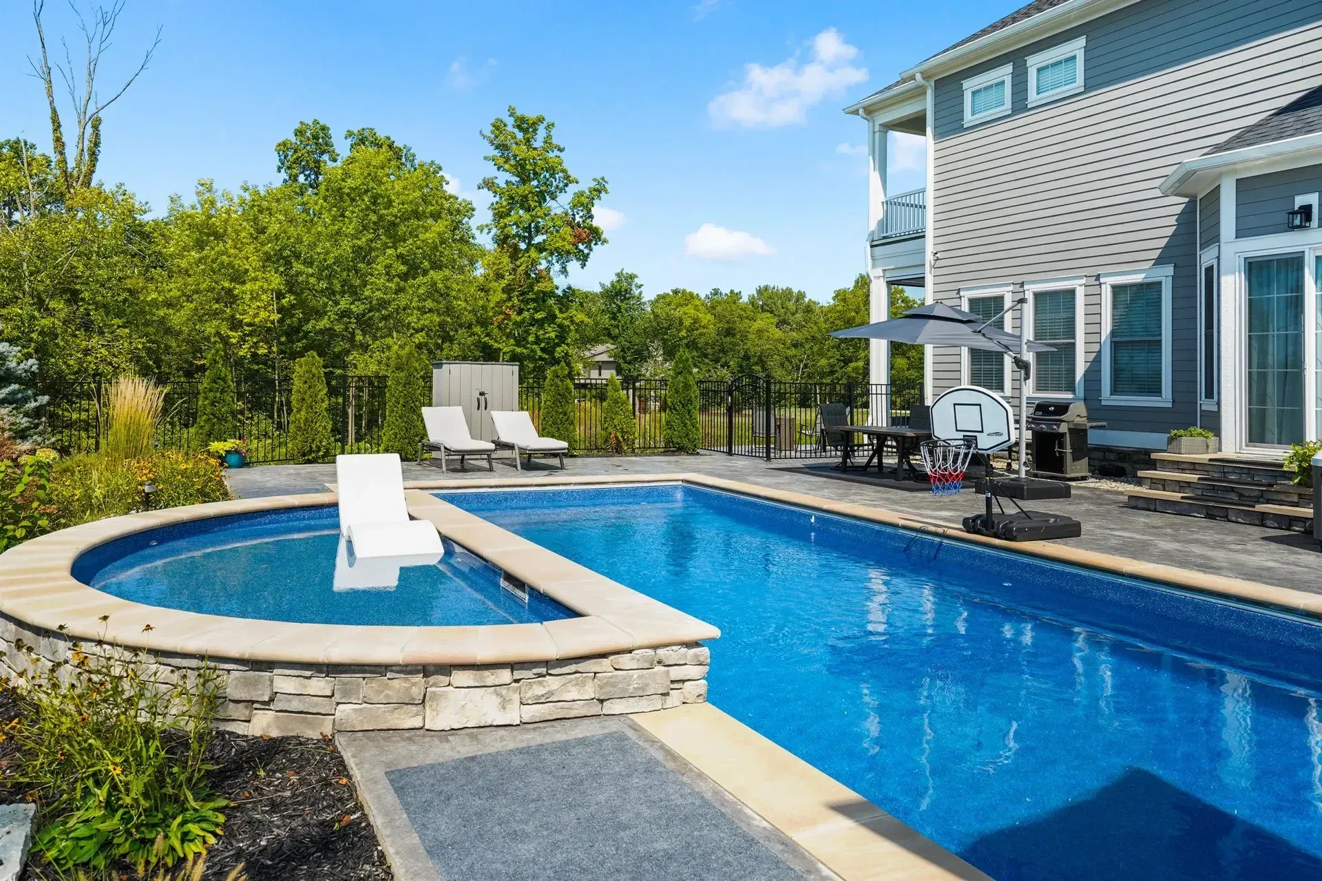 A backyard pool with a connected hot tub. The blue water reflects the sunny sky. A house sits beside the pool.