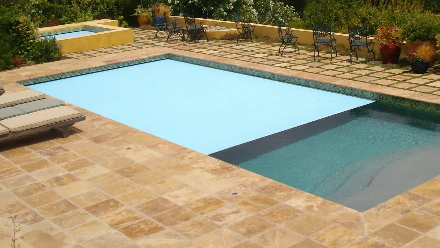 A rectangular swimming pool with a light blue cover partially extended. Beige tile surrounds the pool, and a smaller hot tub is in the background.