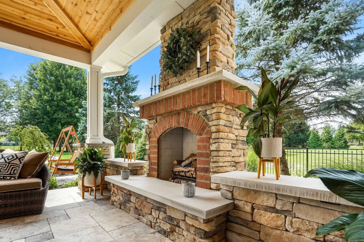 Outdoor patio with a stone fireplace and a wooden ceiling. A seating area is on the left with plants and greenery.