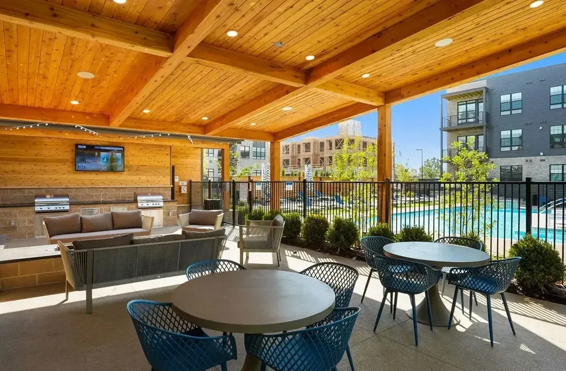 Outdoor covered patio with seating, a pool in the background, and a wooden ceiling. The patio area has tables, chairs, and a built-in sofa.