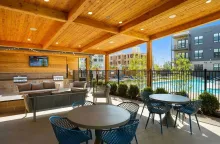 Outdoor covered patio with seating, a pool in the background, and a wooden ceiling. The patio area has tables, chairs, and a built-in sofa.
