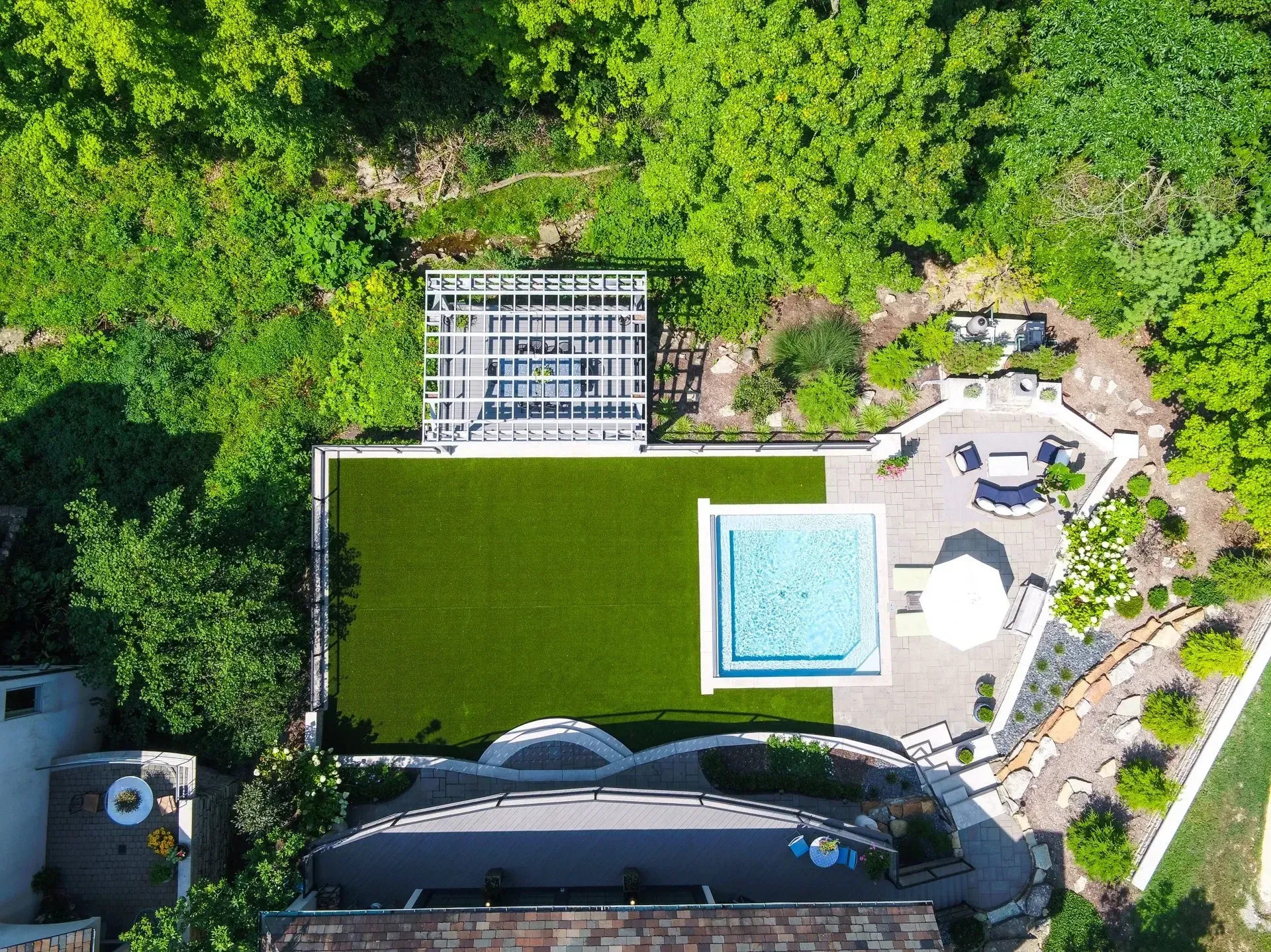 Aerial view of a backyard with a rectangular pool, green lawn, pergola, and patio surrounded by lush greenery.