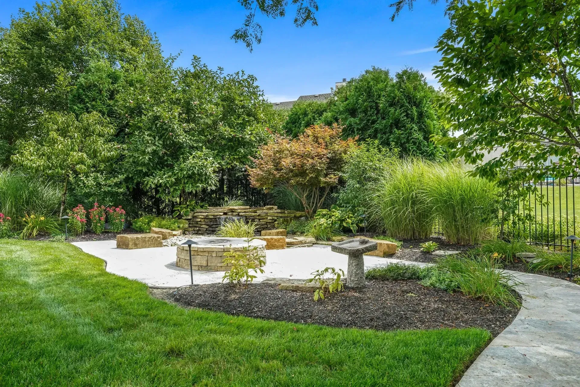 A backyard with a patio, fire pit, and landscaping. Green grass surrounds a stone path and garden beds filled with shrubs and trees.