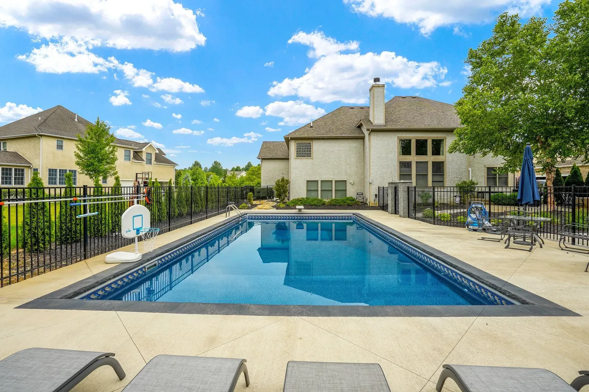 Swimming pool in a backyard with a house and sunny blue sky. Pool has dark tile edge, lounge chairs are visible.