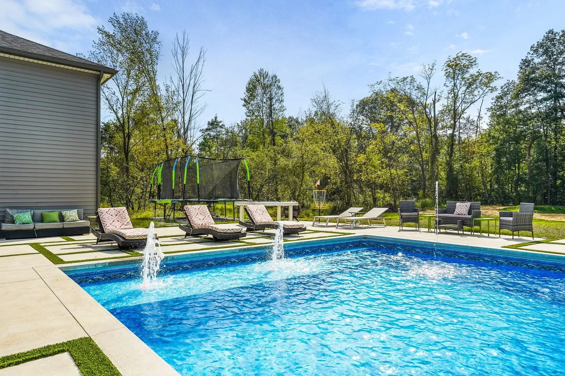 A luxurious backyard pool with fountains and lounge chairs, backed by trees and a house on a sunny day.