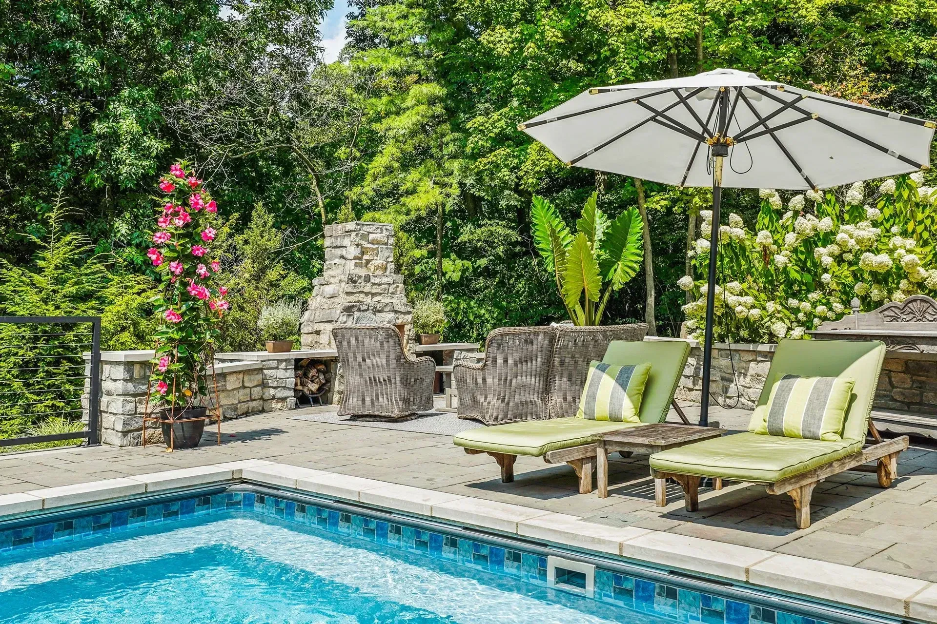 Backyard with a pool, lounge chairs under a sun umbrella, and a stone fireplace. Green foliage surrounds the patio.