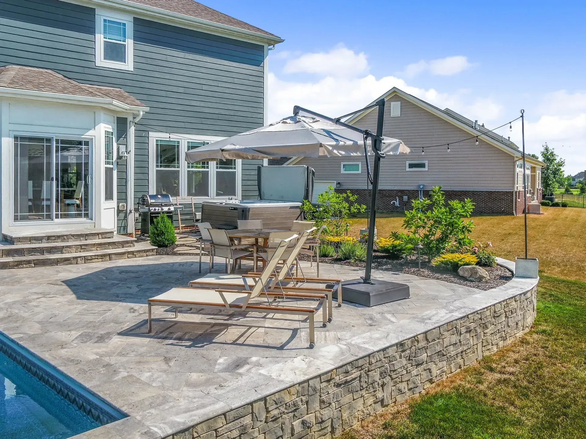 A backyard patio with a pool, lounge chairs, and umbrella. A raised stone wall borders the patio, next to a grassy lawn.