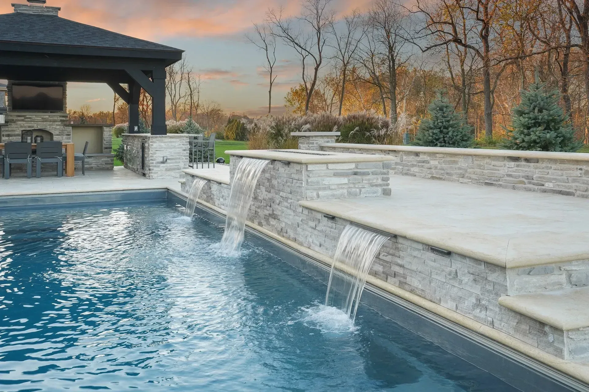 Swimming pool with cascading waterfalls and a gazebo in a backyard setting. Water is blue, structures are stone and beige.