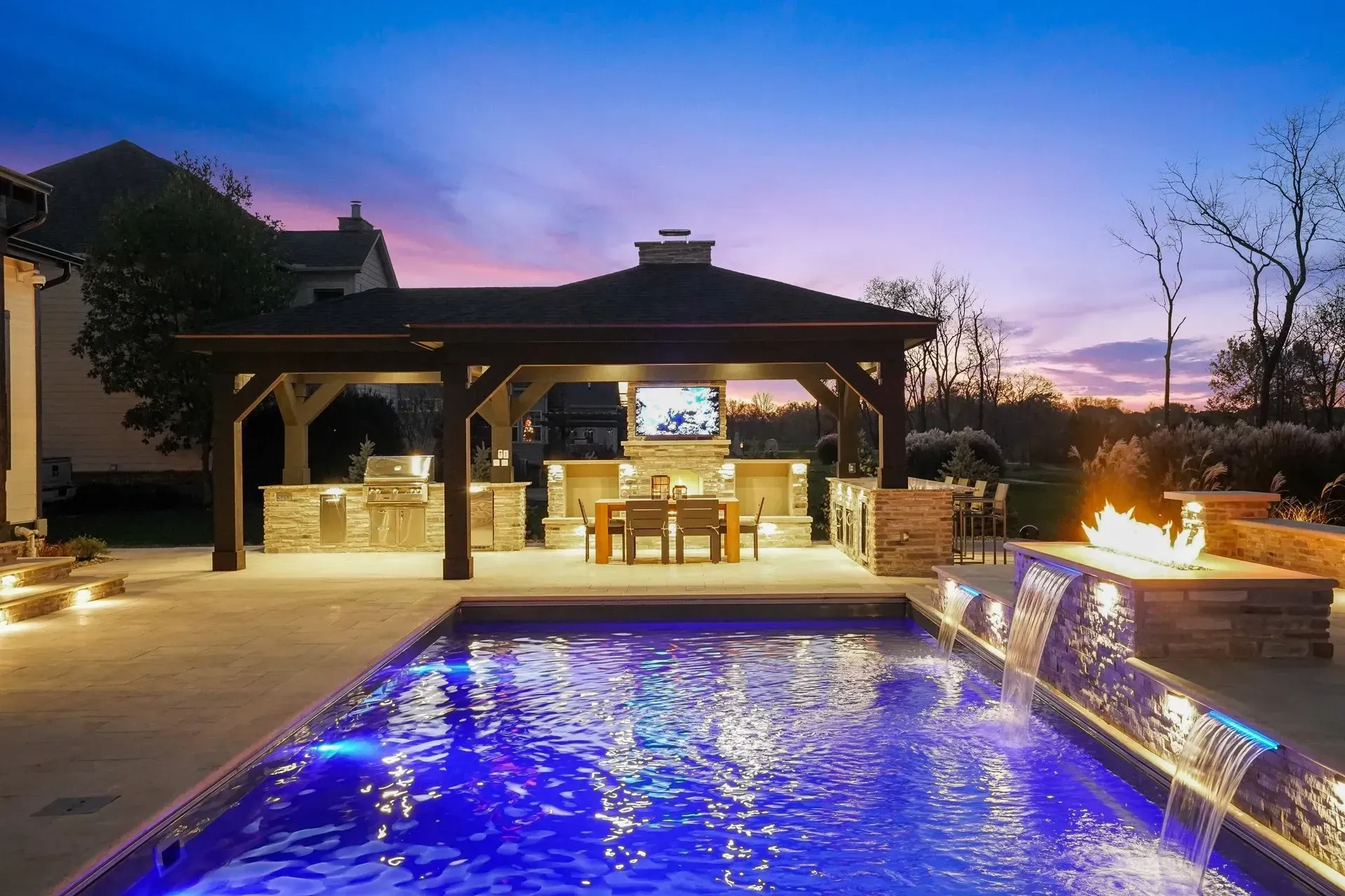 Outdoor kitchen and pool area at dusk with blue-lit water, a gazebo, and a fire pit.