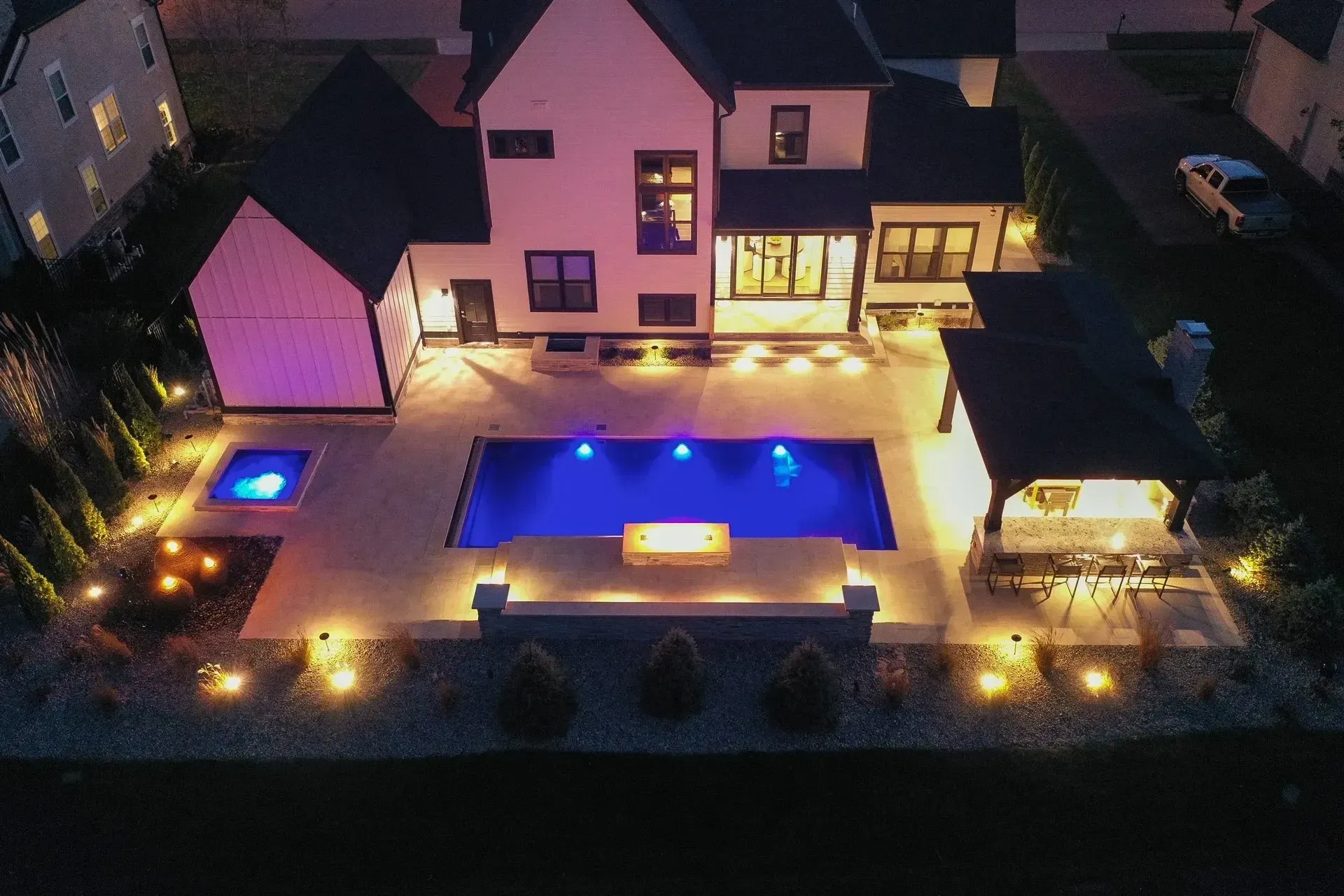 Nighttime aerial view of a modern home with a pool and outdoor entertaining area lit with blue, white, and warm lights.