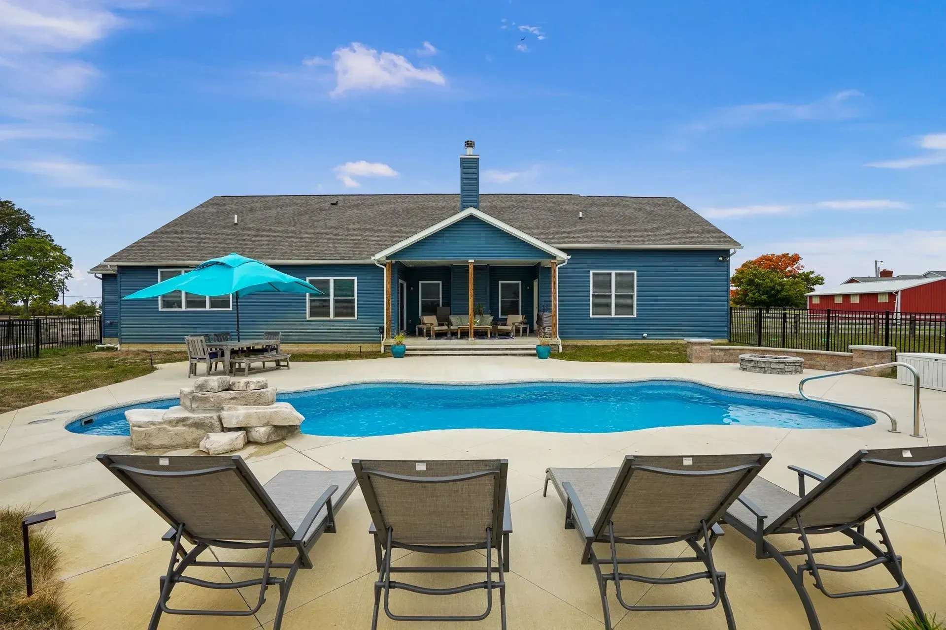 Backyard with a blue house, swimming pool, lounge chairs, and a patio table with an umbrella on a sunny day.
