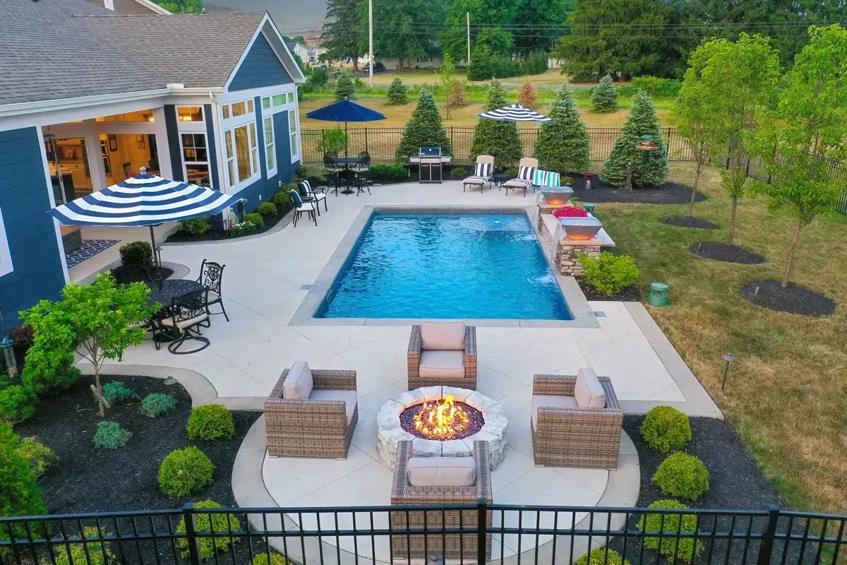 Backyard with a pool, fire pit, seating, and a house. The pool is surrounded by a concrete patio. Blue house with outdoor dining area.