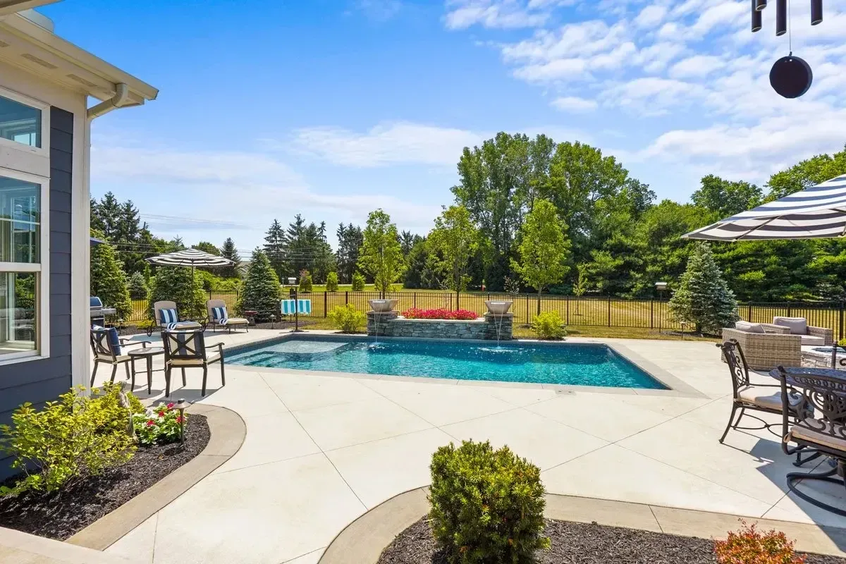 A backyard with a pool, patio furniture, and landscaping. The scene is bright and sunny, with a blue sky and green trees.