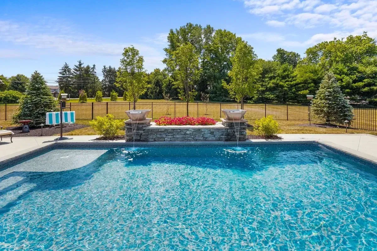 Pool with a stone wall feature, red flowers, and trees under a blue sky.