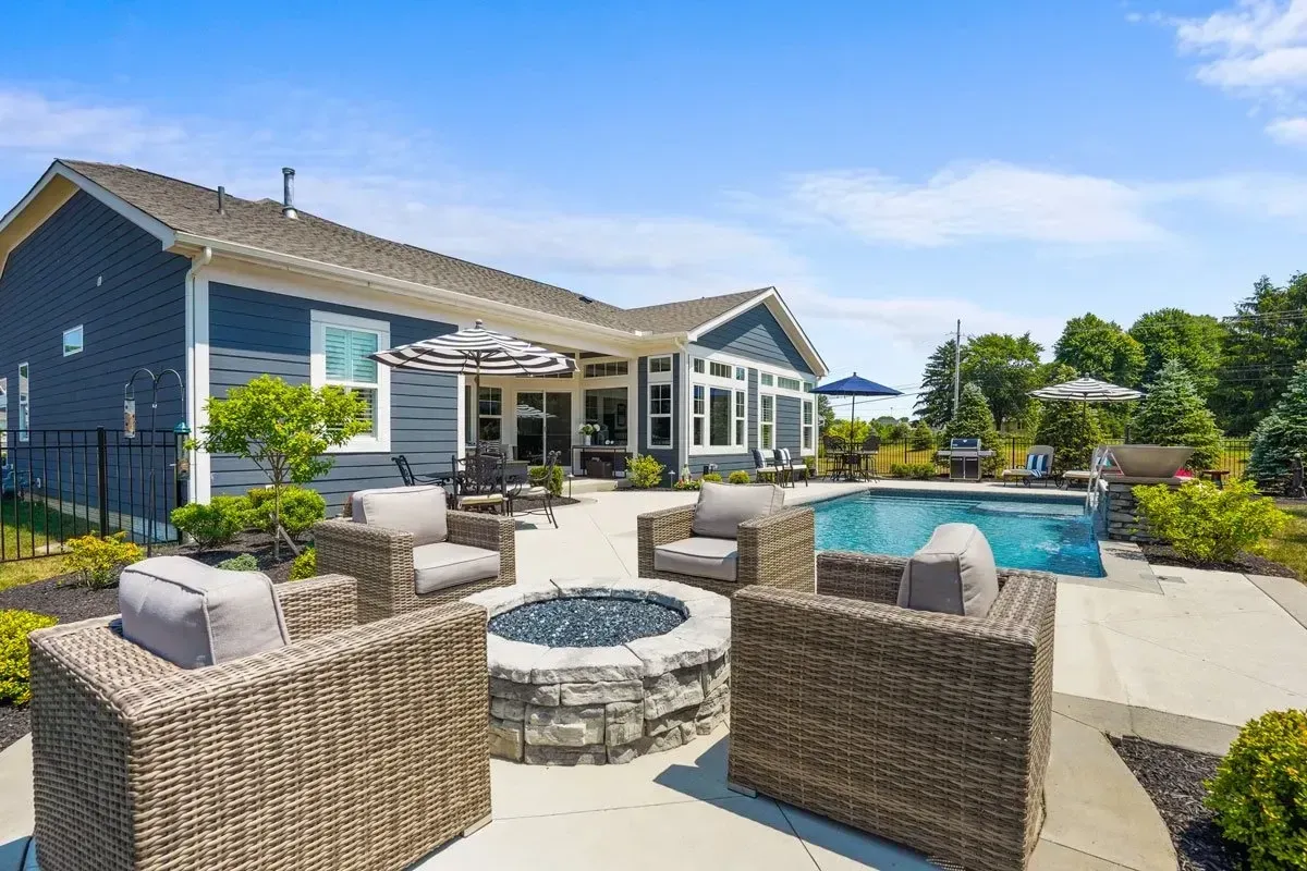 Backyard patio with seating area, fire pit, and swimming pool. Blue house in the background on a sunny day.