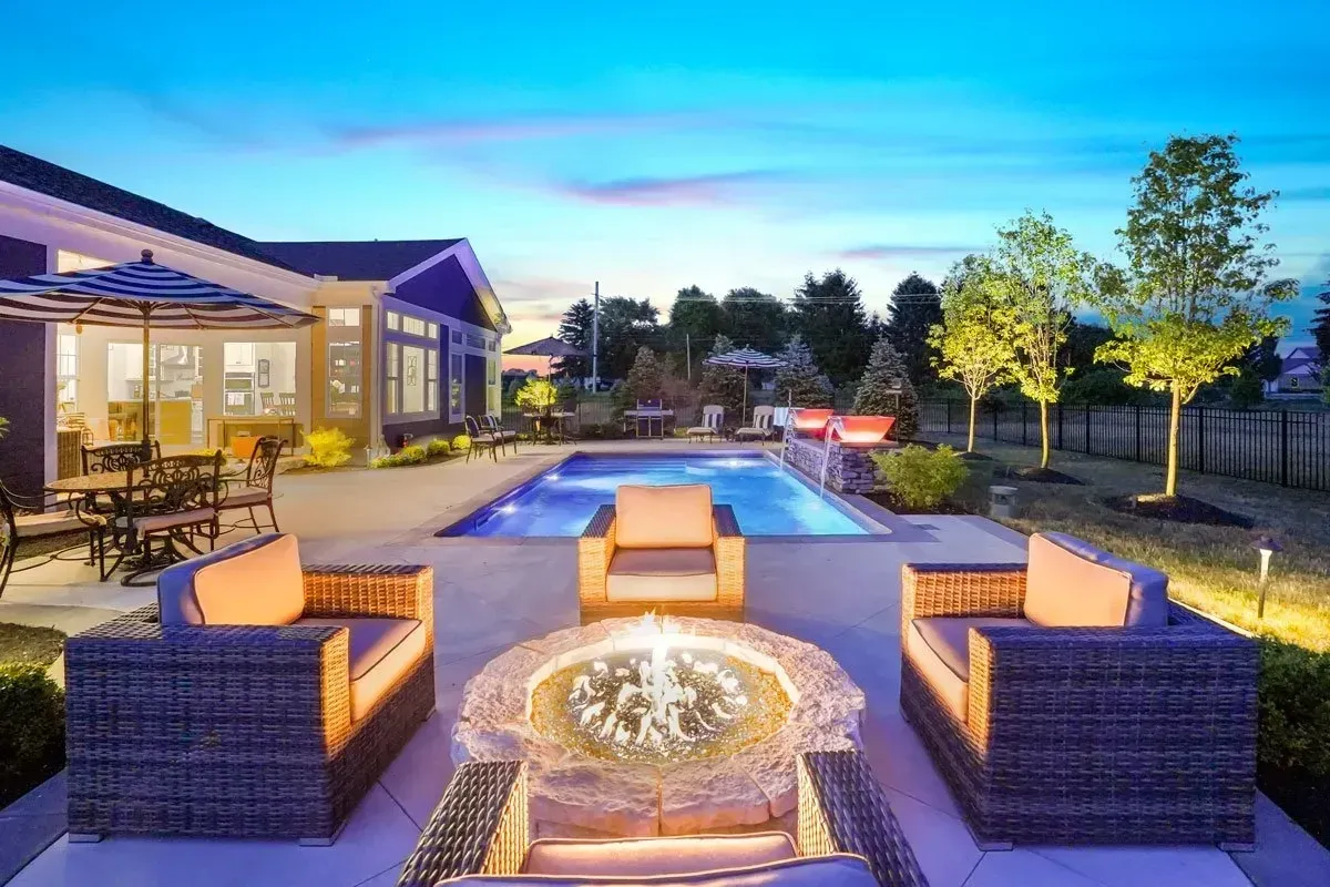 Outdoor patio with a fire pit, seating, pool, and house at dusk.