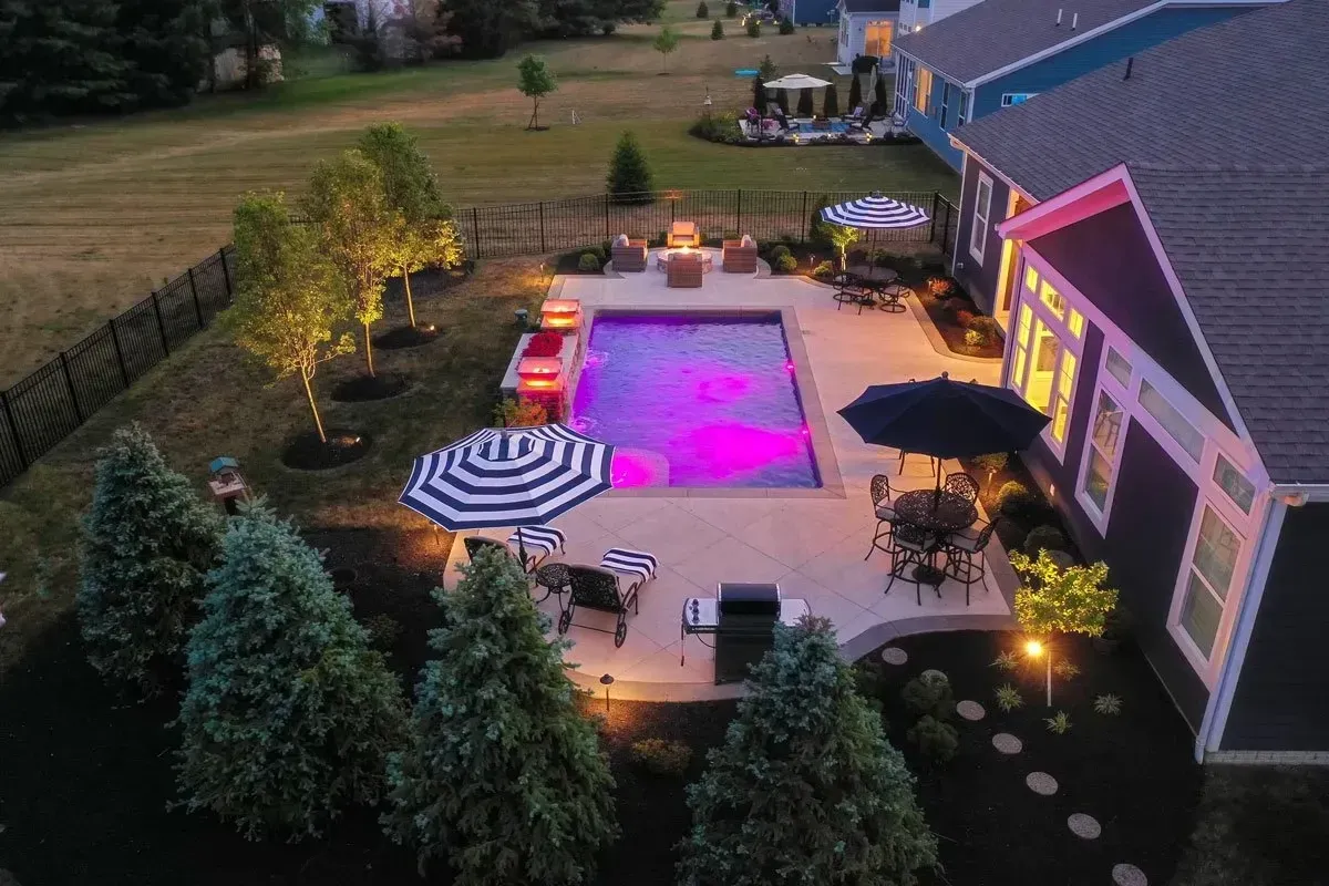 Aerial view of a backyard pool area at dusk. The rectangular pool glows purple. Outdoor furniture and umbrellas are set up on the patio.