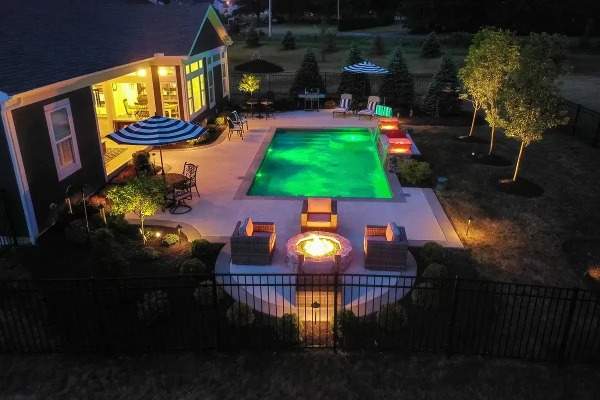 Nighttime aerial view of a backyard with a lit green pool, patio, fire pit, and house. Warm lights illuminate the scene.