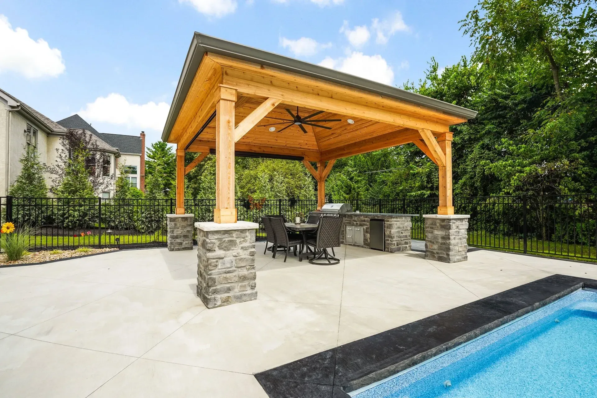 Outdoor kitchen with wooden pergola, concrete patio, pool, and stone pillars. Dining set and grill under the shelter.