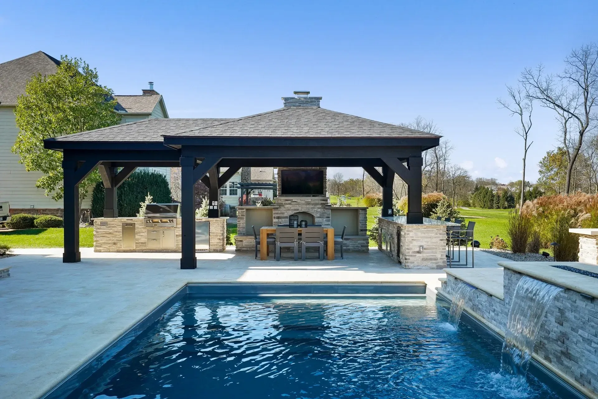A stone fireplace on a covered patio with potted plants and a wooden ceiling, set against a backdrop of trees.