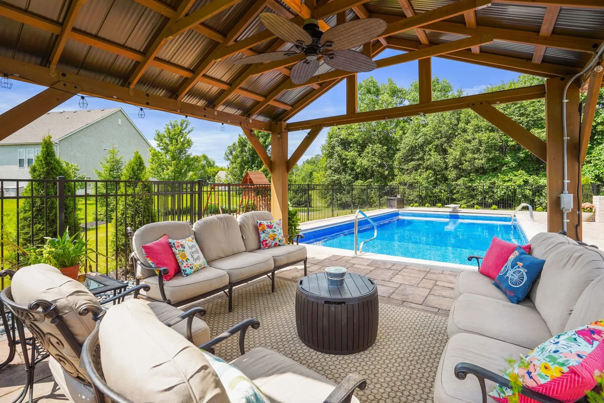 Outdoor patio with a pool view. Beige couches with colorful pillows under a wooden gazebo, pool in the background.