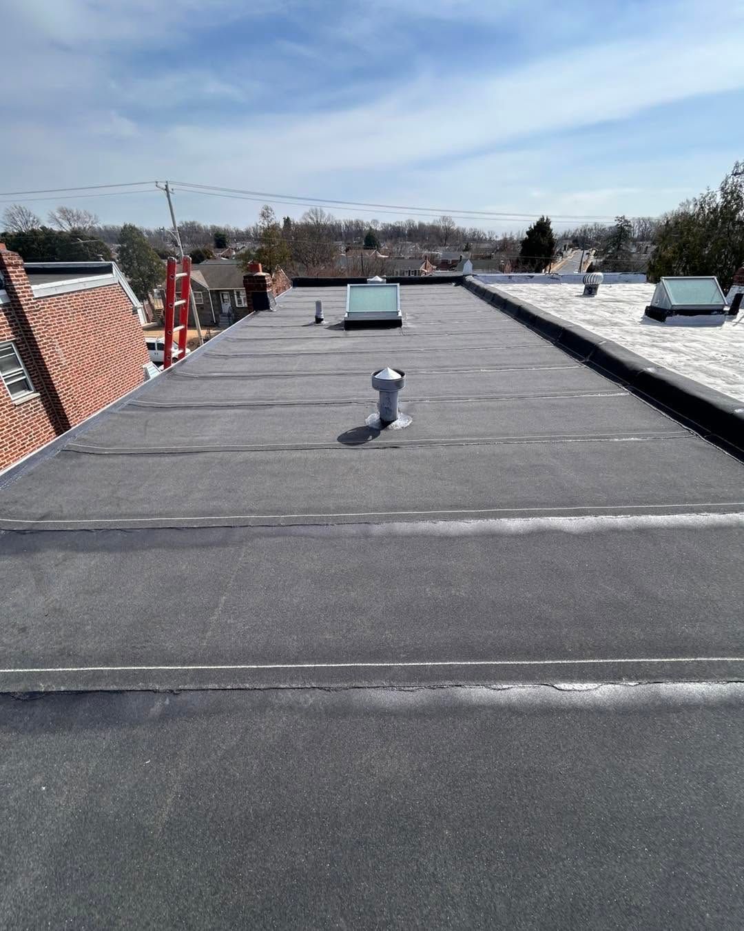 Flat black rooftop with vents and brick parapet under a blue sky, overlooking distant houses