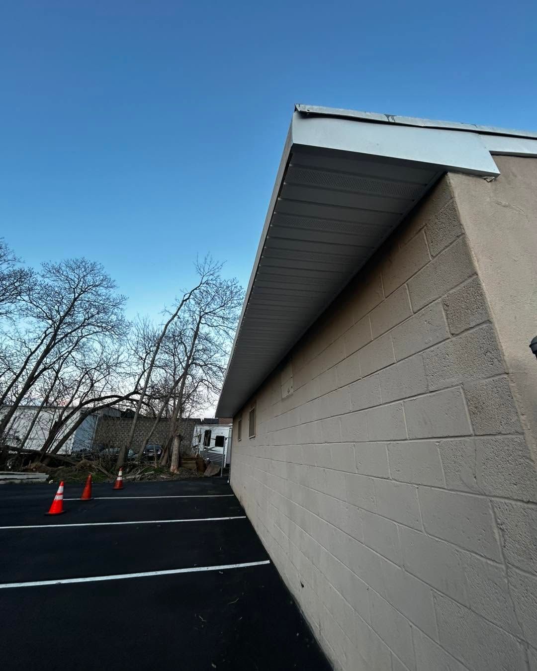 Side view of a beige building with a dark roof overhang beside an empty parking lot and trees.
