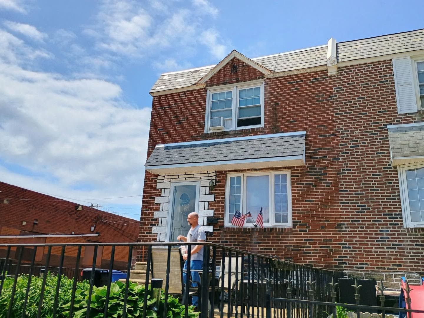 Person standing by the front steps of a brick house with a black fence and blue sky.