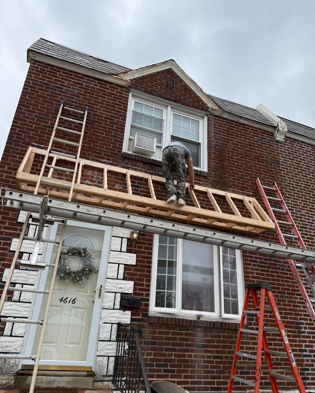 Worker on scaffolding repairing a brick house facade with white trim and a front door.
