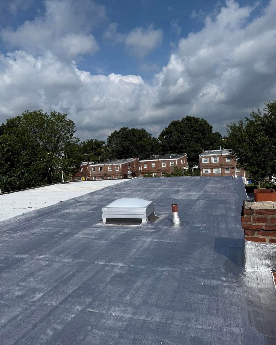 Flat rooftop with a white vent, overlooking brick buildings and trees under a cloudy sky.