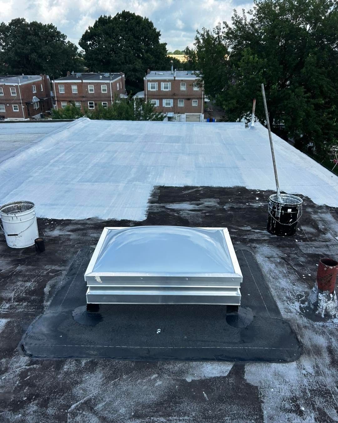 Rooftop with a white skylight hatch on a dark patch, surrounded by vents and distant brick buildings.