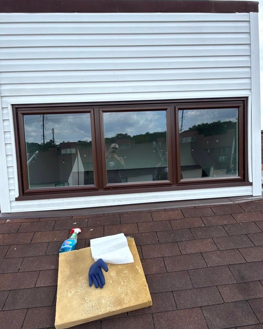 Roof work setup with gloves, spray bottle, and cloth on shingles in front of a window.