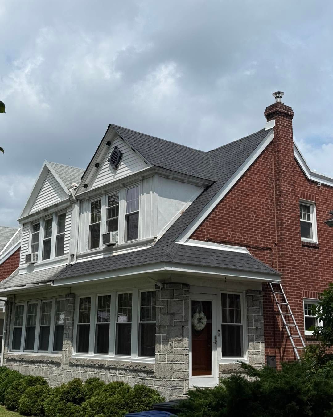 White two-story house with red brick chimney, dark roof, and stone lower facade under a cloudy sky