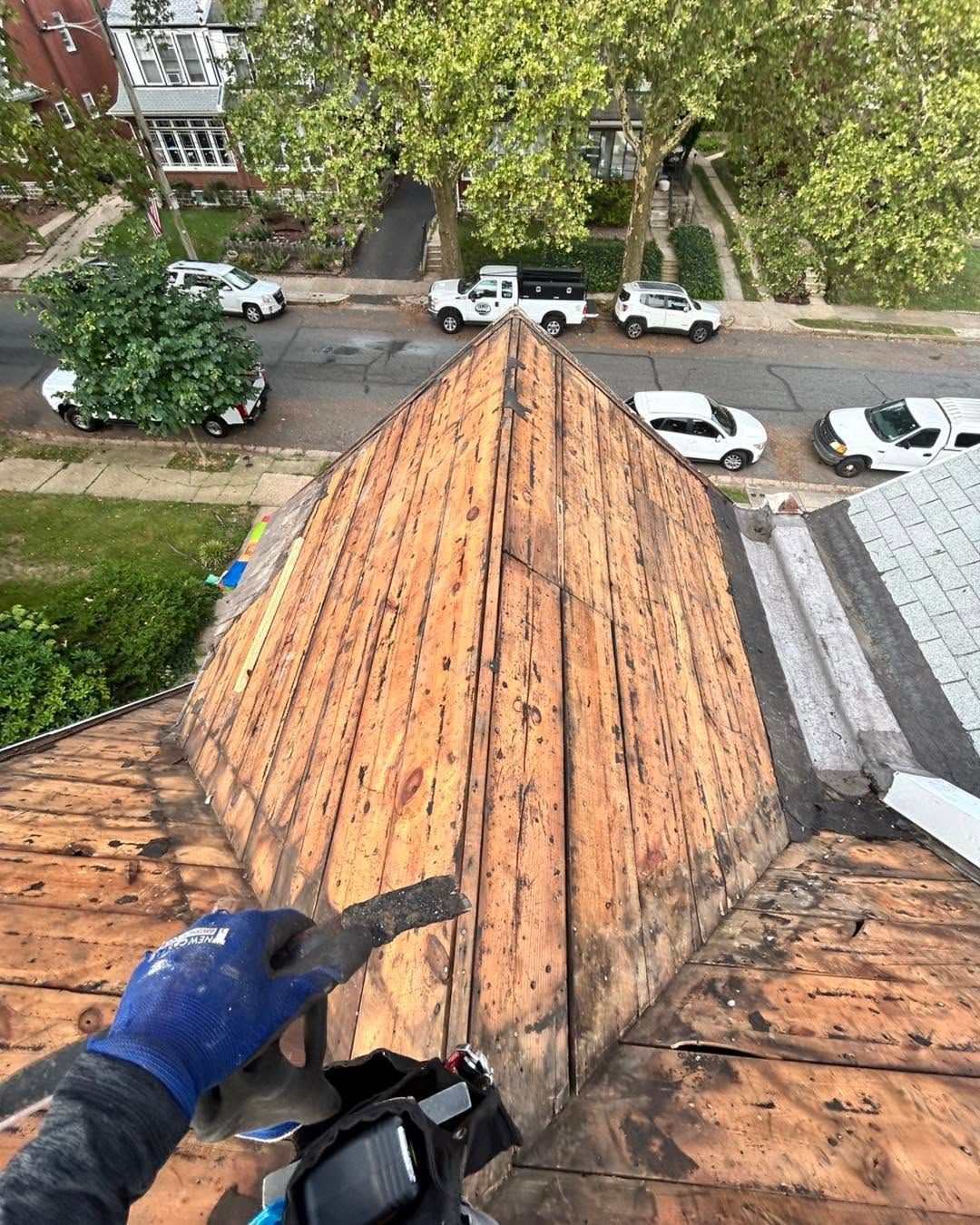 Worker on a roof inspecting orange shingles, looking down at a street below.